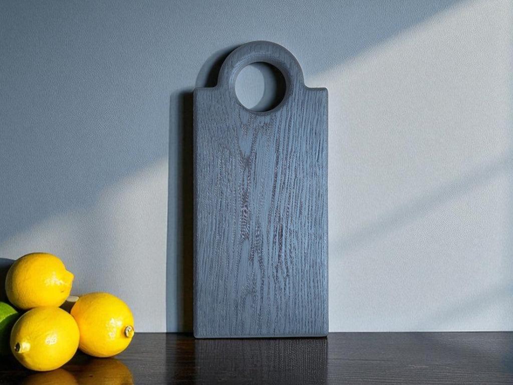  Schneidebrett "Oldrye Black Wide Loop" neben frischen Zitronen auf dunklem Holztisch, im Hintergrund eine graue Wand mit Lichtschatten.