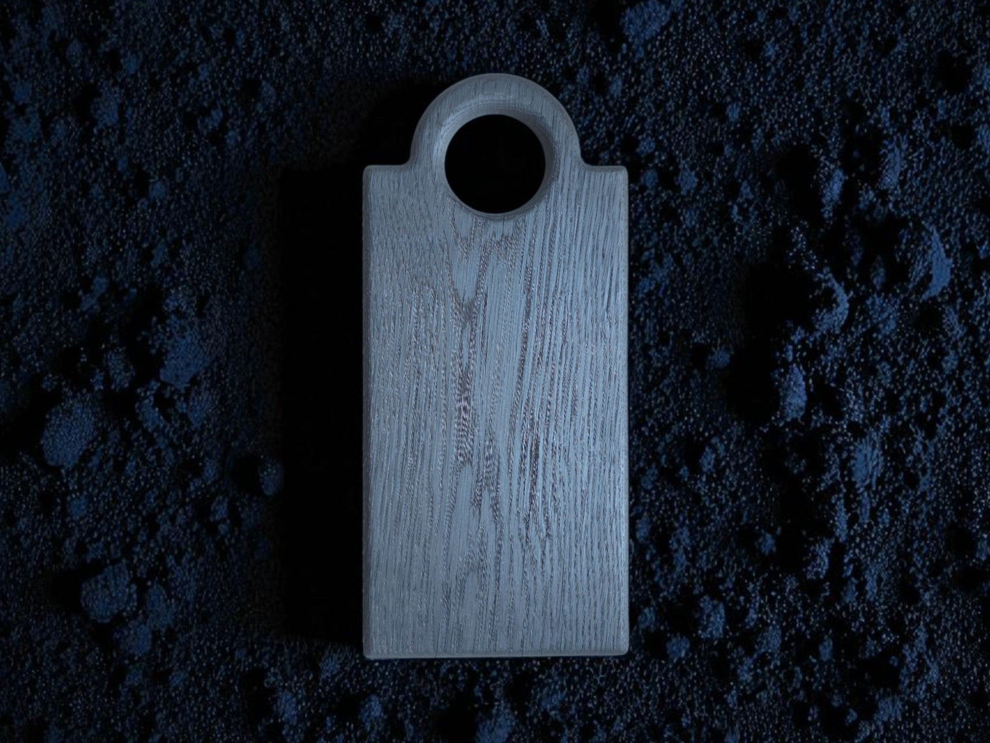 Das Schneidebrett "Oldrye Black Wide Loop" liegt auf einem Bett aus dunklem, blaugrauem Pulver, das die Oberfläche umschließt.