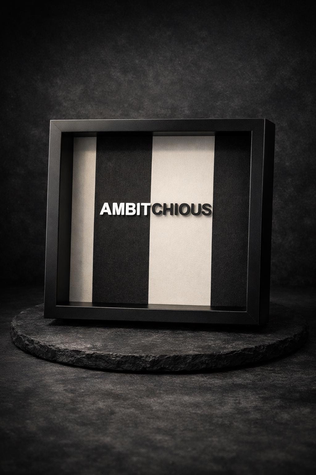 FACTIONARY™ Statement Frame „AMBITCHIOUS“ von OLDRYE®, schwarzer tiefer Rahmen mit erhabener 3D-Typografie auf schwarz-weiß gestreiftem Textilstoff, modernes Wortkunst-Design im Format 25x25 cm