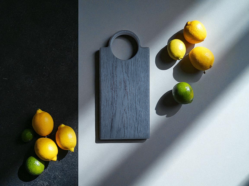 Eine stilvolle Draufsicht auf das handgefertigte "Slim Loop" Servierbrett aus Yakisugi-Eichenholz. Das minimalistische, graue Brett liegt auf einer zweifarbigen Oberfläche und wird von hellen Limetten und Zitronen umrahmt. Die natürlichen Früchte und der Schattenwurf betonen die einzigartige Haptik und die natürliche Schönheit des Boards.